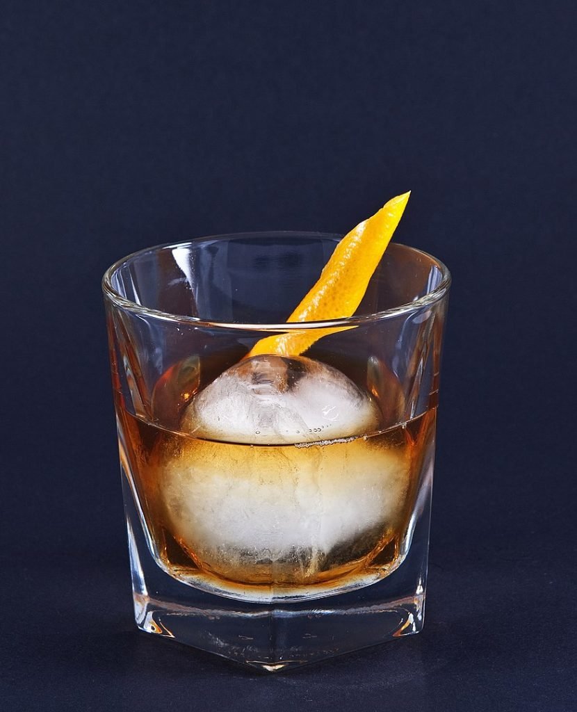 A Whiskey Old Fashioned on an iceball, decorated with an orange twist.