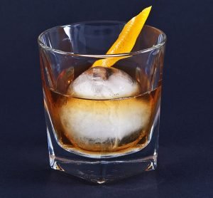 A Whiskey Old Fashioned on an iceball, decorated with an orange twist.